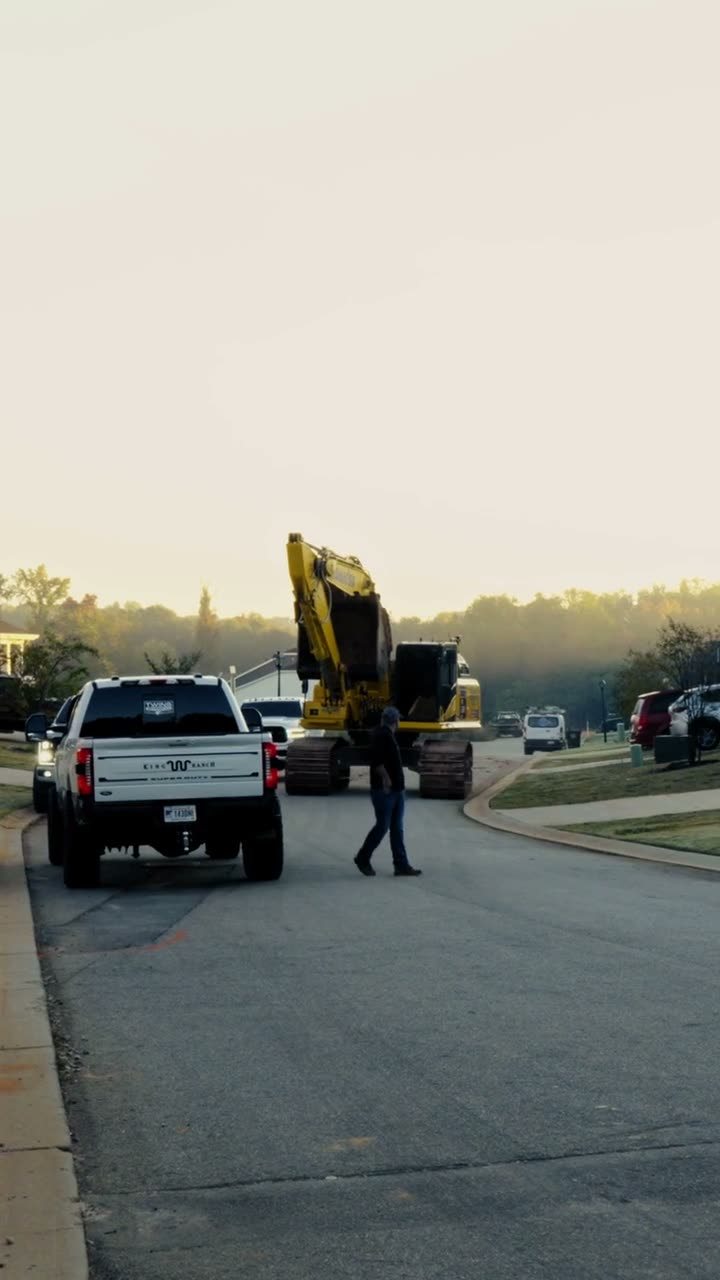 Pickup truck leading an excavator on a lowboy trailer down a neighborhood street